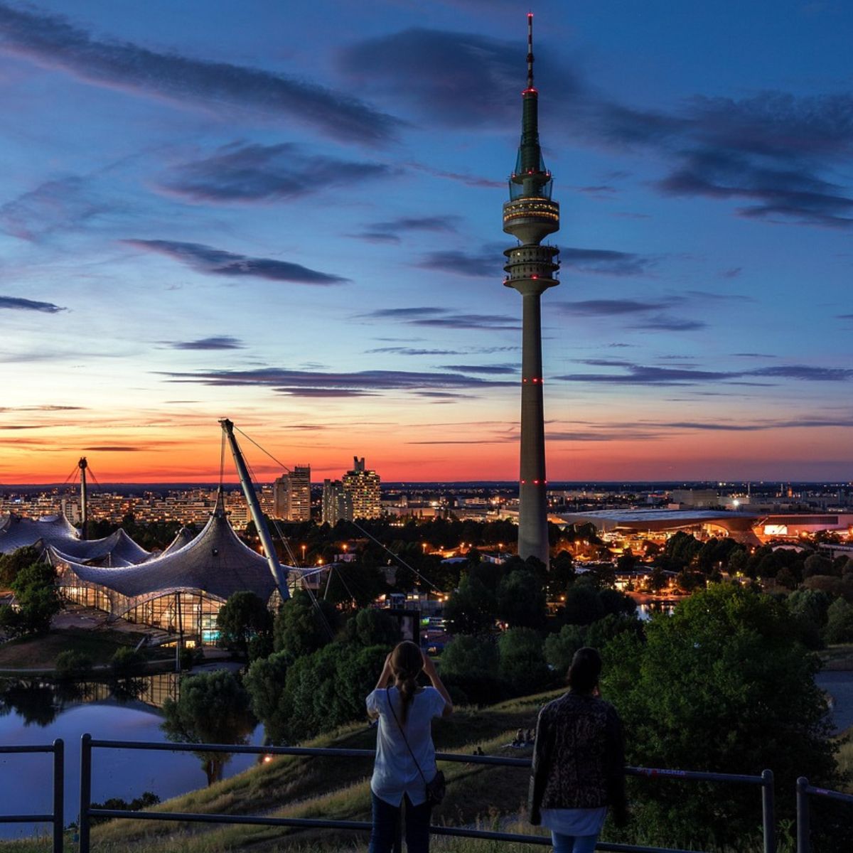 Blick auf den München Olympia Park mit Olympiaturm bei Sonnenuntergang und zwei Personen im Vordergrund.