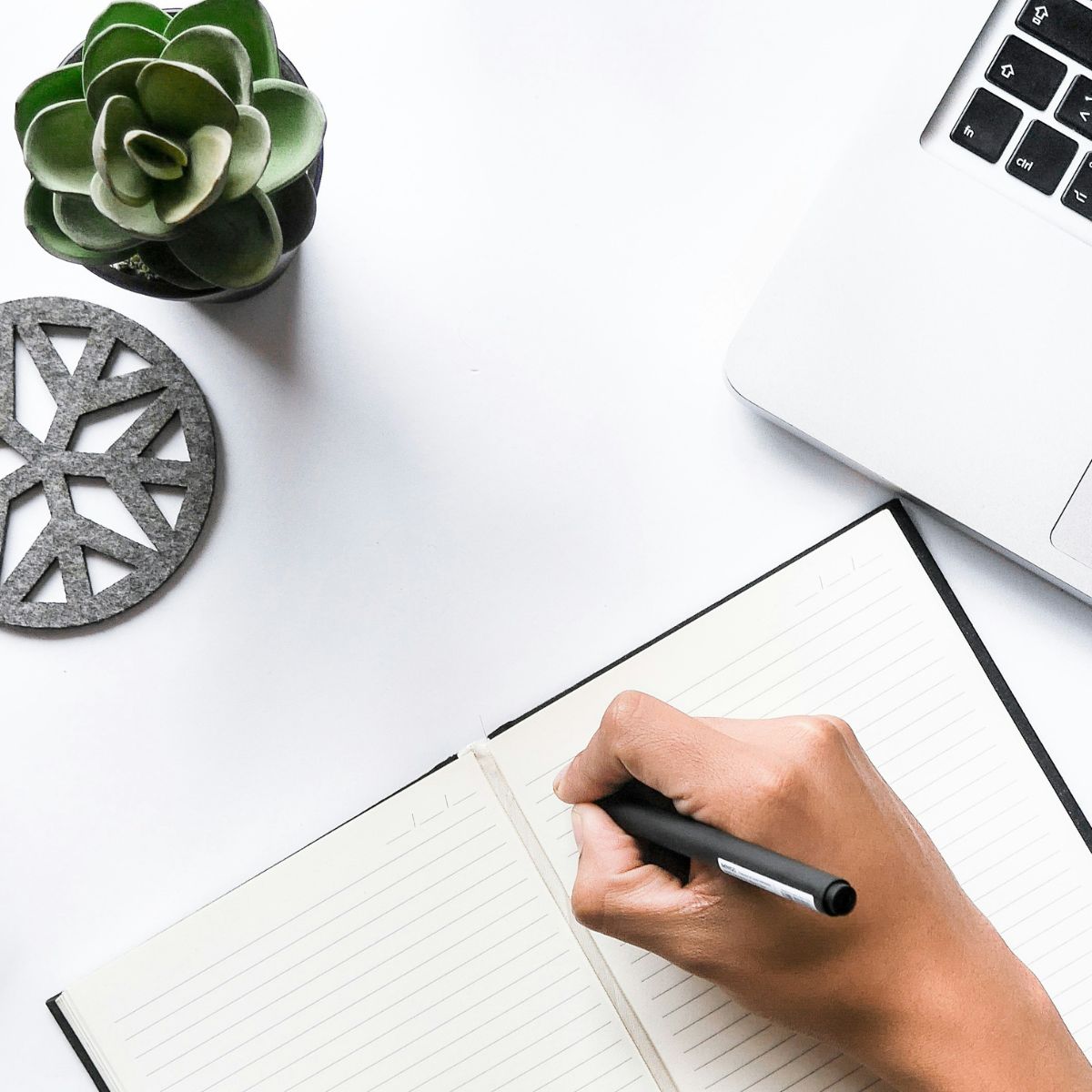 and schreibt in ein Notizbuch neben Laptop und Pflanze – Sinnbild für konzentriertes Schreiben & Lernen.