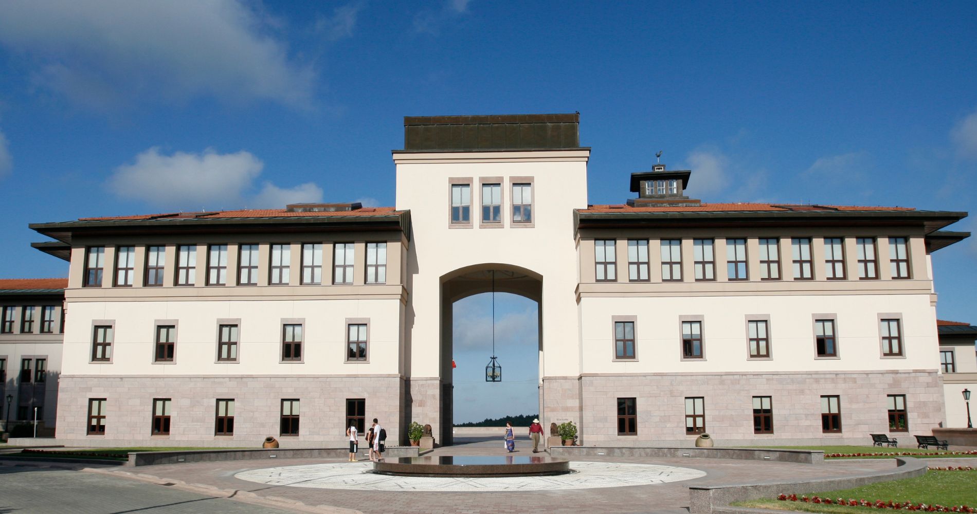 Koç University – Repräsentatives Hauptgebäude mit großem Torbogen, Brunnen und blauem Himmel.