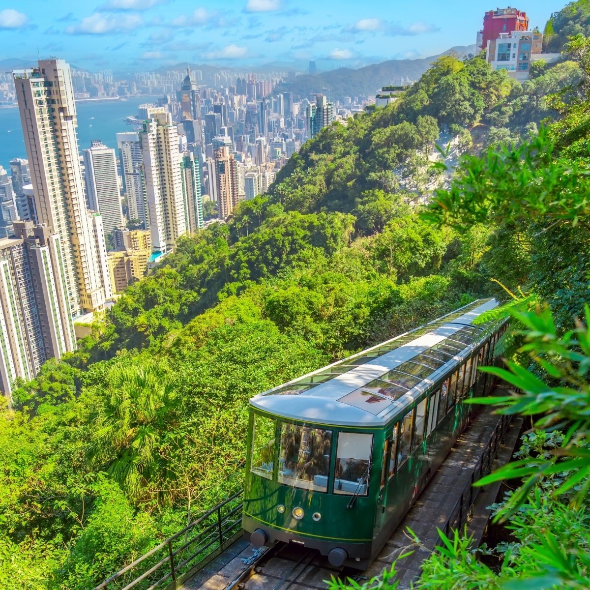 Hong Kong – Grüne Peak Tram fährt den Victoria Peak hinauf, im Hintergrund die Skyline und der Hafen.