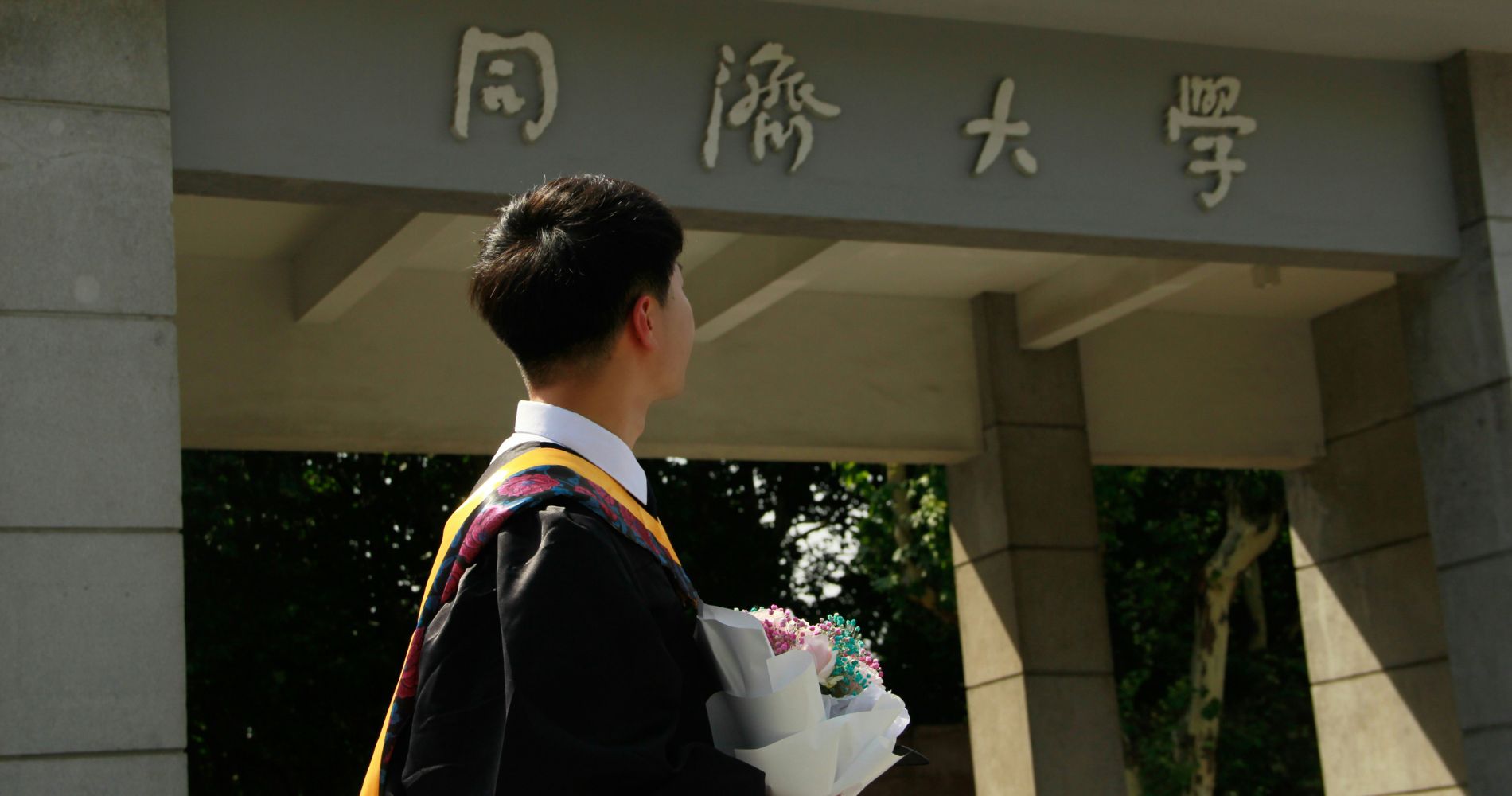 Tongji University – Absolvent im Talar mit Blumenstrauß vor dem chinesischen Schriftzeichen-Eingangstor.