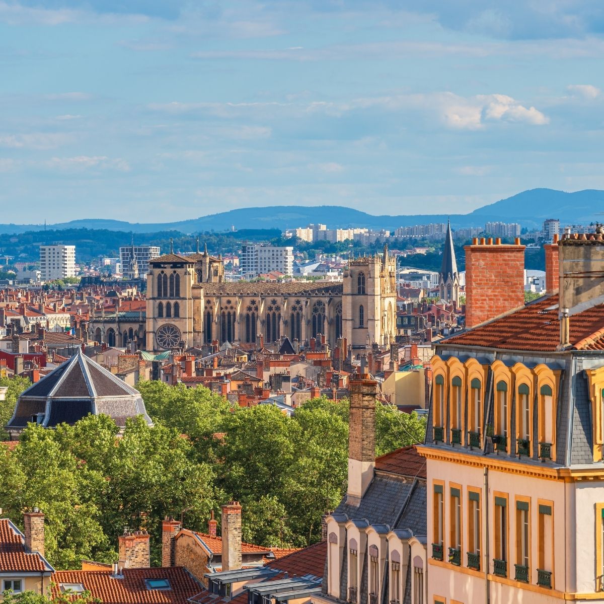 Lyon – Stadtpanorama mit gotischer Cathédrale Saint-Jean und roten Ziegeldächern vor grünen Hügeln.
