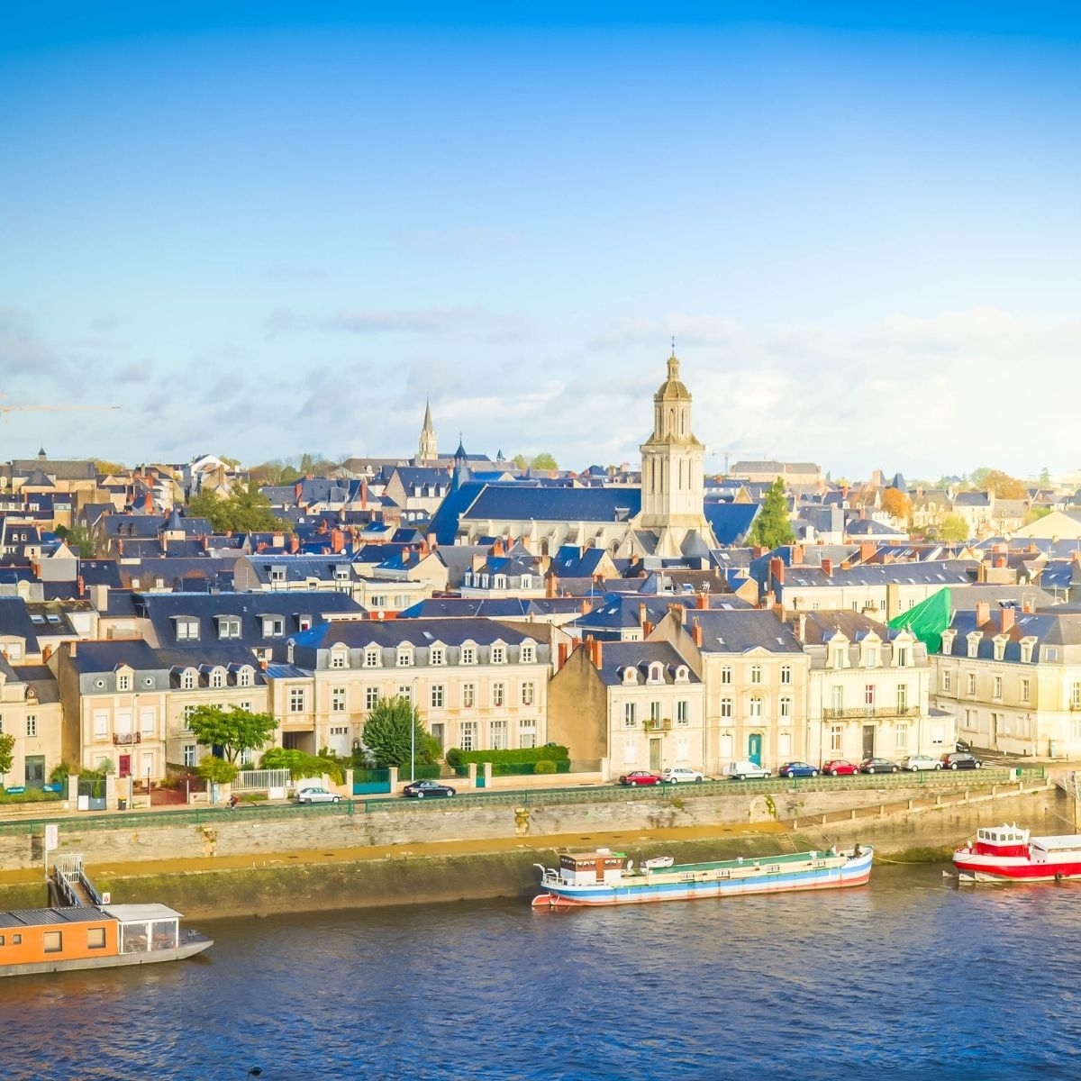 Angers – Stadtpanorama mit Kirchturm und historischen Häusern am Ufer der Maine mit Booten im Vordergrund.