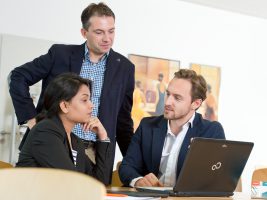 Three professionals in suits/DBA candiates sitting in front of a laptop discussing a topic.