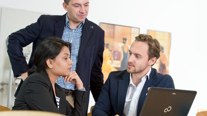 Three professionals in suits/DBA candiates sitting in front of a laptop discussing a topic.