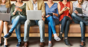 People side by side on a bench using different digital devices such as a laptop, smartphone or tablet.