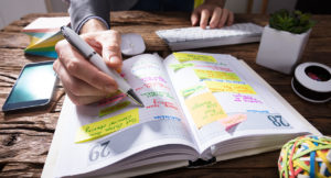 Lifelong Learning: Close-up Of A Businessperson's Hand Writing Schedule In Diary With Pen On Wooden Desk