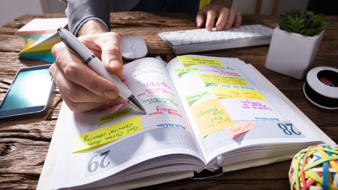 Lifelong Learning: Close-up Of A Businessperson's Hand Writing Schedule In Diary With Pen On Wooden Desk