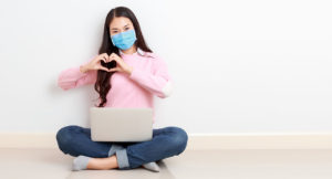 Young Asian woman wearing surgical mask sits cross legged with a laptop on her legs on the floor and forms a heart with her hands