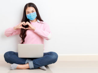 Young Asian woman wearing surgical mask sits cross legged with a laptop on her legs on the floor and forms a heart with her hands