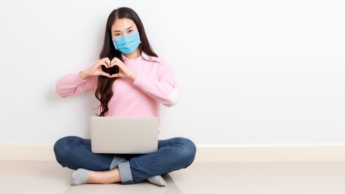 Young Asian woman wearing surgical mask sits cross legged with a laptop on her legs on the floor and forms a heart with her hands