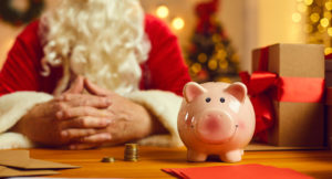 Close-up of piggy bank on desk with presents and Christmas charity event invitations with old Santa Claus in blurred background.