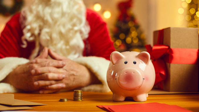 Close-up of piggy bank on desk with presents and Christmas charity event invitations with old Santa Claus in blurred background.