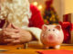 Close-up of piggy bank on desk with presents and Christmas charity event invitations with old Santa Claus in blurred background.