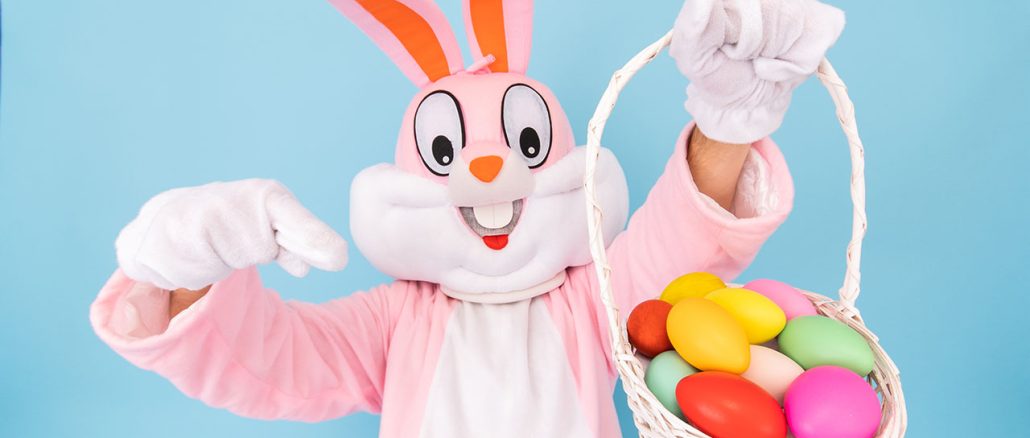 Easter Bunny pointing at a basket with colored eggs he is holding in his hand.