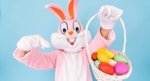 Easter Bunny pointing at a basket with colored eggs he is holding in his hand.