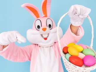 Easter Bunny pointing at a basket with colored eggs he is holding in his hand.