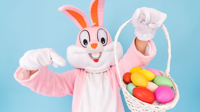 Easter Bunny pointing at a basket with colored eggs he is holding in his hand.