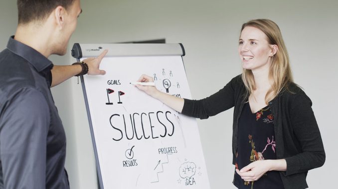 Two students from Munich Business School standing together at a flipchart with "success" written on it, representing a typical practical work situation at private universities.